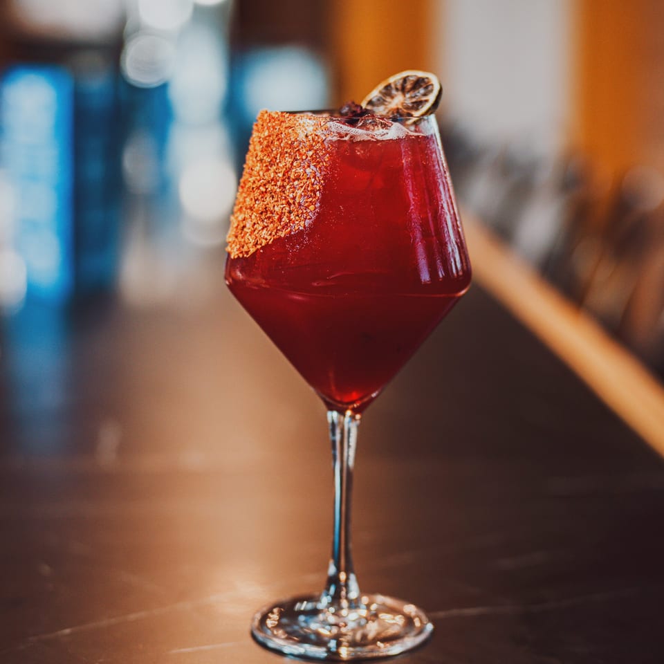 Red cocktail in a stemmed glass with a chili-salt rim and dried citrus garnish on a warmly lit bar.