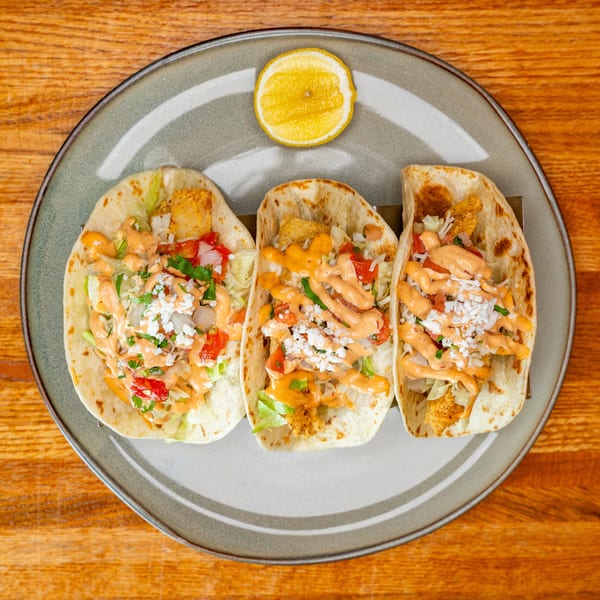 Three crispy tacos topped with lettuce, tomato, sauce, and cheese, served with a lemon.