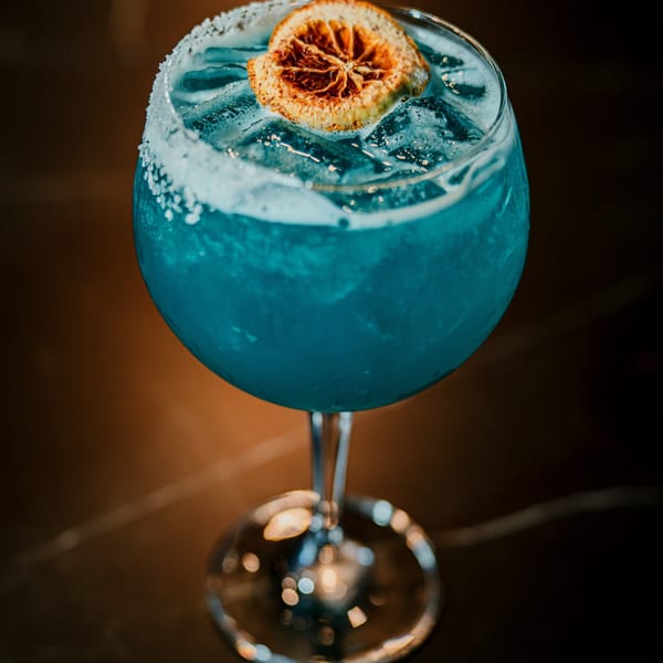 Blue cocktail with a salted rim, ice, and a dried orange slice garnish on a dark table.