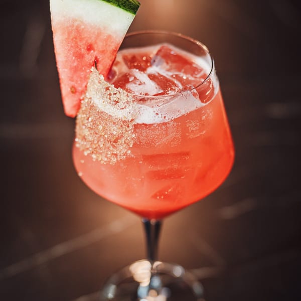 Bright red cocktail with ice, a sugar-rimmed glass, and a watermelon wedge garnish against a dark background.
