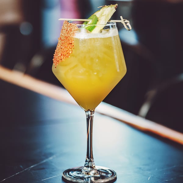 A vibrant yellow cocktail in a stemmed glass, garnished with pineapple and a chili-salt rim.
