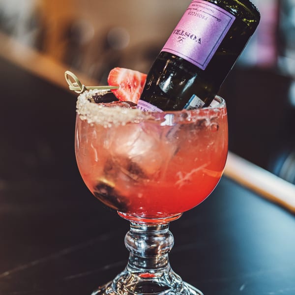 A red cocktail with ice, a strawberry garnish, and a small bottle of La Vostra Prosecco.