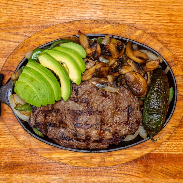 Sizzling steak with avocado, mushrooms, onions, and a green pepper in a cast iron skillet on wood.