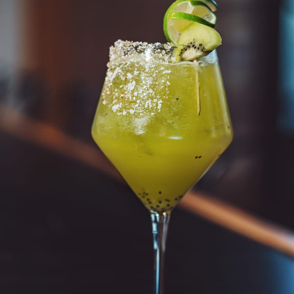 Green cocktail in a sugar-rimmed martini glass garnished with lime slices and kiwi against a dark background.
